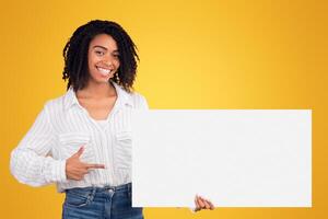 African Woman Holding Blank Paper On Yellow Background, Smiling And Pointing At It, Empty Space photo