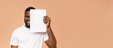 African American young man with a short beard and a white t-shirt is smiling while holding a blank white piece of paper in front of his face. He is standing in front of peach-colored background. photo