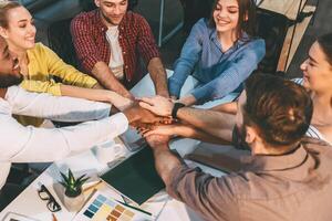 A group of diverse coworkers enthusiastically join hands over a desk, symbolizing teamwork and unity in a modern office environment. photo