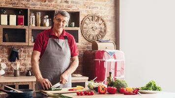A man, wearing glasses and an apron, is smiling as he cuts vegetables on a cutting board in a modern kitchen. He is surrounded by a variety of colorful vegetables photo