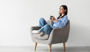 Positive young lady in casual wear sitting in armchair with crossed legs, warming hands on cup of hot coffee, enjoying warm drink against white studio wall, copy space photo