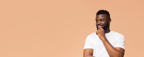 A young black man with a beard is smiling and looking off to the side with his hand on his chin. He is wearing a white t-shirt and is standing in front of a light brown background, copy space photo