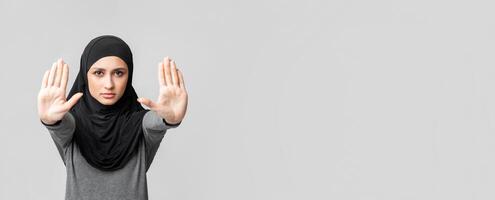 No discrimination. Concerned arabic woman in headscarf showing stop sign gesture with two hands, demonstrating palms over grey background with free space photo