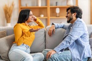 Indian man and woman are seated on a beige couch, engaged in conversation. Both individuals are facing each other, with the woman gesturing with her hands as they talk. photo