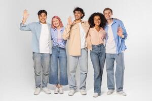 Five young adults, including three men and two women, stand together in a studio setting, smiling and waving to the camera. They are all wearing casual clothes and appear to be happy photo