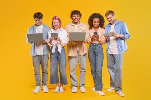 Five young adults, standing in a row in front of a yellow background, are using different forms of technology, including laptops, tablets, and smartphones. photo
