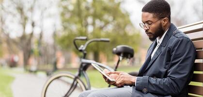 An African American man is on a park bench, working on a digital tablet with a bike nearby. The setting is calm and green, reflecting a mix of business and leisure in nature. photo