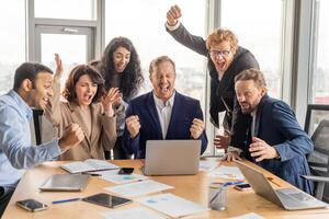 A diverse group of business professionals is gathered around a conference table in a modern office, celebrating a success. They are all smiling and cheering, with their hands raised in the air. photo