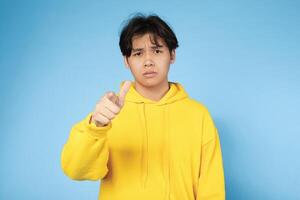 Asian young guy wearing a bright yellow hoodie stands against a blue wall. He is pointing forward and looking directly at the camera with a serious expression. photo