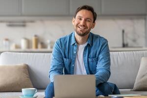 A man is sitting on a couch in his living room, smiling and looking at the camera while using his laptop. There is a cup of coffee on a table in front of him. photo