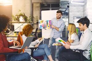 A group of professionals is gathered for a meeting in a contemporary office setting, reviewing and discussing ideas. Man stands confidently in front of a whiteboard covered with colorful sticky notes photo