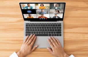 A top-down view of a persons hands using a laptop for a conference. The laptop screen shows a grid of nine tiles, each featuring a different person. photo