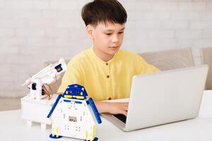 A young boy is sitting at a desk, using a laptop to program a small robot. The robot is made of white plastic with blue accents and has a robotic arm. The boy is focused on the screen photo