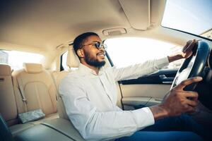 African American man with a beard and sunglasses drives a car while wearing a white shirt. He appears to be enjoying the drive, looking out the window and smiling. photo