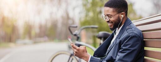 Black business professional is on his phone, sitting on a park bench with a bicycle next to him. The scene is calm and modern, blending work and leisure in a natural setting. photo