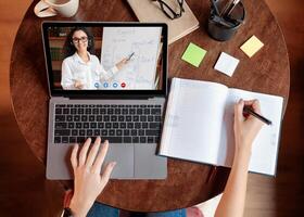 Distance Education Concept. Above top view of female student sitting at desk, writing in notebook, taking notes, using laptop, having online class with teacher. Remote studying from home, quarantine photo