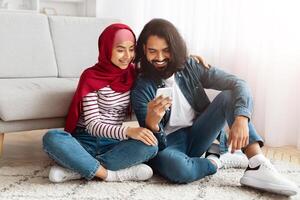 Eastern man and woman are seated on the floor, both engrossed in their cell phone screen, focused as they browse content together, sharing an intimate moment in their modern digital interaction. photo