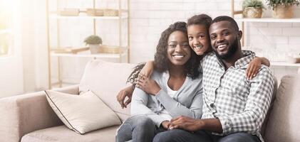 Family portrait. Happy african american parents and their daughter embracing and posing to camera while sitting together on couch at home photo