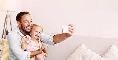 Selfie With Daughter. Smiling dad taking photo and hugging his toddler girl sitting on sofa, spending time together