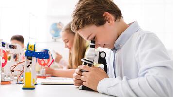 A young boy, wearing a white shirt, looks intently through a microscope, focusing on a slide. He is sitting at a desk in a classroom setting, surrounded by other students and educational materials photo
