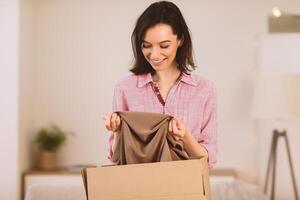 Moving Concept. Excited girl getting ready for moving out, packing clothes in paper box, copyspace photo