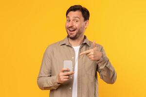 A man with a beard is holding a smartphone in his left hand while smiling and pointing at it with his right hand. He is standing against a bright yellow background. photo