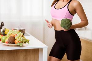 A woman wearing workout clothes holds a head of broccoli in her kitchen. A bowl of fruit is on the counter in the background. photo