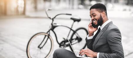 Black man professional is using his laptop and talking on his phone, enjoying an outdoor workspace with his bicycle next to him. The setting is dynamic and relaxed, capturing a modern work lifestyle. photo