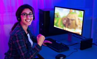 A young woman sits in front of a computer screen, wearing a headset and smiling as she plays a game. The computer monitor is illuminated by neon pink and blue lighting photo