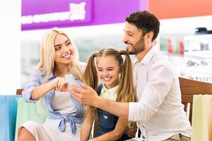 A young family, a father, a mother, and their young daughter, are sitting together in a shopping mall. They are looking at a smartphone together and smiling photo