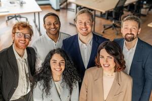 Five diverse business professionals stand together in a modern office, looking directly at the camera and smiling. They are wearing business casual attire, high angle view photo