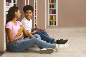 sonriente negro Adolescente Pareja sentado en piso a estante para libros en biblioteca y mirando a cada otro, panorama con gratis espacio foto