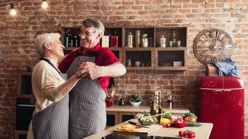 A senior couple in aprons, smiling and holding each other, are dancing in a kitchen. Fresh vegetables are on the counter in front of them. photo