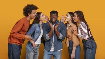 A group of five young friends stand in front of a yellow background. Two women whisper to the man in the center of the group, while the two other friends lean in to listen to what is being said photo
