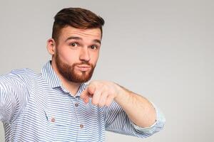 You are cool. Young bearded guy making selfie and pointing you with finger against grey studio background photo