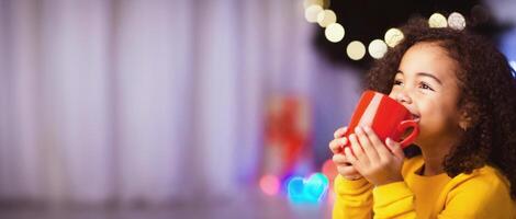 Cute little african girl with cup of hot cocoa sitting near Christmas tree, empty space photo