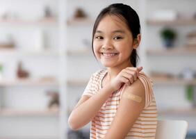 Healthy asian kid girl got vaccinated, sitting on couch at home and showing adhesive band on her shoulder, smiling at camera, panorama with copy space. Kids vaccination, immunization concept photo