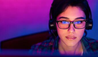 A woman wearing glasses and headphones is looking intently at a computer screen. The room is lit in a purple and blue color scheme, creating a vibrant and modern atmosphere photo