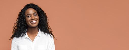 Black woman smiling warmly at the camera. She is wearing a white shirt and her dark curly hair is pulled back from her face. The background is a simple tan color photo