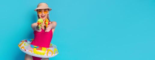 This image shows a woman dressed in a pink swimsuit and a straw hat, holding two water guns in front of her. She is smiling and looking at the camera. Behind her is a light blue background. photo