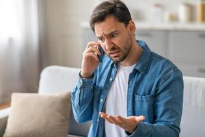 A man with a concerned expression sits on a sofa in a living room, talking on a mobile phone. He appears frustrated, with one hand raised in the air, as he speaks. photo