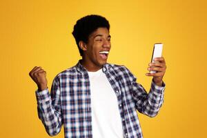 Winner Concept. African Guy Using Smartphone over Yellow Studio Background photo
