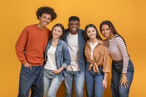 Group of five diverse young adults standing in front of a yellow background. They are all smiling and looking at the camera. The group appears to be friends and is enjoying each others company. photo