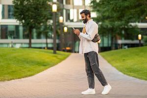 Indian man is walking down a sidewalk while engrossed in his cell phone, his attention focused on the screen rather than his surroundings, multitasking as he navigates the urban environment. photo