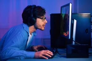 A young man with headphones and glasses is intensely focused on his computer screen, playing a game. The room is dimly lit with blue and purple hues, creating a mood of late-night gaming photo