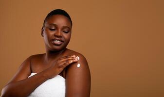 Daily Skincare Routine. Portrait of young nude African American woman wrapped in white towel applying body lotion on shoulder after shower, brown studio background, banner, panorama, free copy space photo