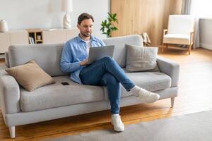 A man is seated on a couch, focused on his laptop screen. He appears engaged in his work or activities online, with a serious expression. The room is simple, with no other visible people photo