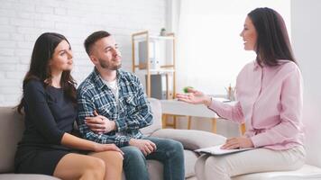 A young couple sits on a couch while a therapist listens intently and speaks to them. The couple appears to be listening and engaged in the conversation. The setting is a modern office photo