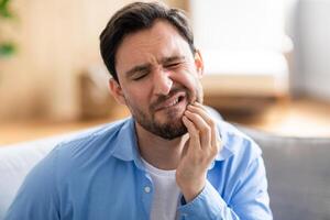 A man is shown sitting on a couch, visibly in discomfort, holding his jaw with a pained expression due to a toothache. He appears to be seeking relief or contemplating seeking dental help. photo