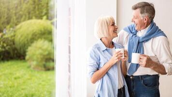 Happy senior couple talking near window and drinking tea, copy space photo